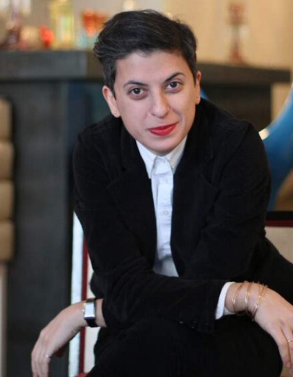 Samira's colour photo: a Middle Eastern woman with short hair and bright red lipstick sits on a stool. She is leaning forward and her arms are crossed upon her knee.