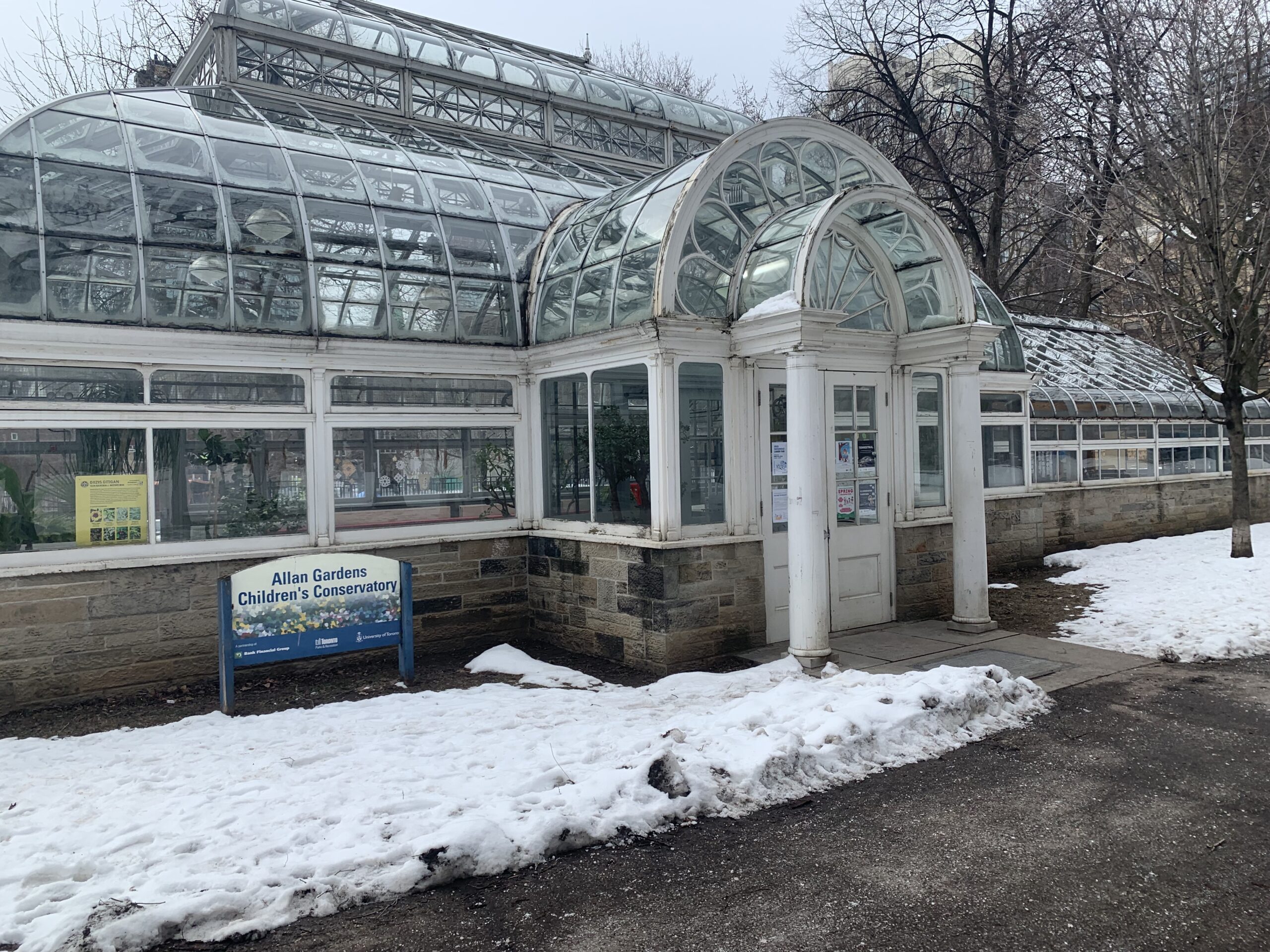 Image of the entrance to the Children's Conservatory, a Victorian-era greenhouse with an ornate entrance made of glass with columns, and a blue sign to the left of the main entrance.