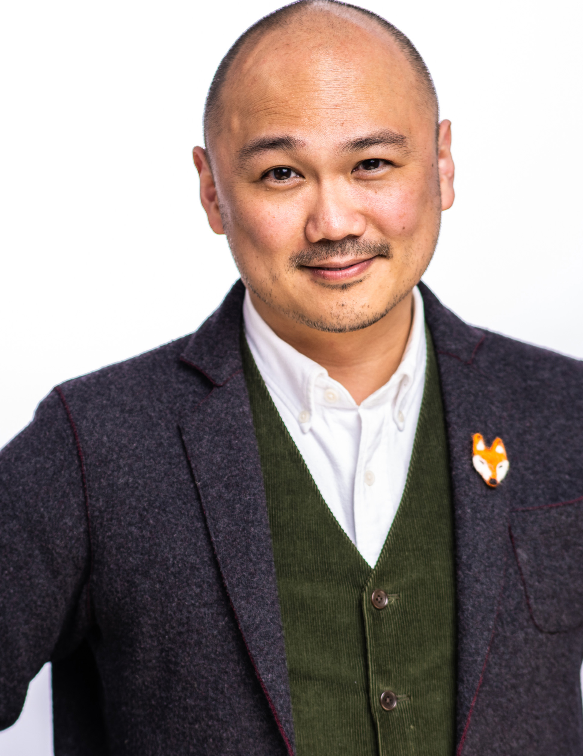 Headshot of Derek Kwan. He is an East Asian man in his 40s, with a shaved head and stubble moustache and beard. He wears a white shirt, a dark green corduroy vest, and a dark grey suit jacket adorned with a needle-felted pin of a fox head.