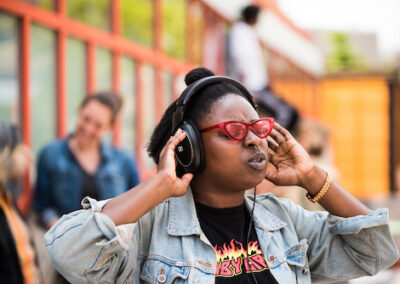 A black woman with short hear and red heart -shaped vintage sunglasses listens with headphones on.