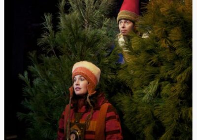 A Woman in Red Winterwear stands in front of Christmas trees while a young man peeks through them