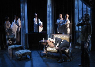 A light illuminates a single chair in on a dark stage. A Maid in a classic black and white outfit lies asleep on it. The rest of the ensemble carry a pillow each as they look at her.