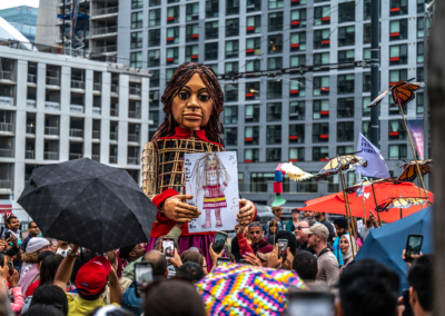 Amal holds a large portrait drawn by an admirer, surrounded by a crowd holding umbrellas, butterfly puppets, and other puppets. In the background are tall buildings.