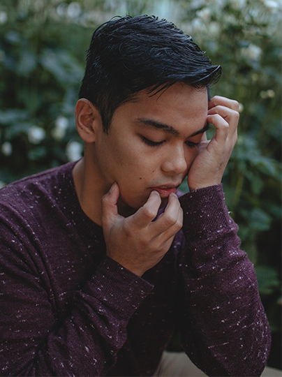 A side profile of Miggy, who crouches amidst bushes and white flowers that recede blurrily into the background. His fingers gently crawl up from his long-sleeved maroon shirt, over his chin and lips, and toward his black, wavy hair. The brown skin of his cheek is caressed by his palm as he looks down with eyes closed in contemplation.