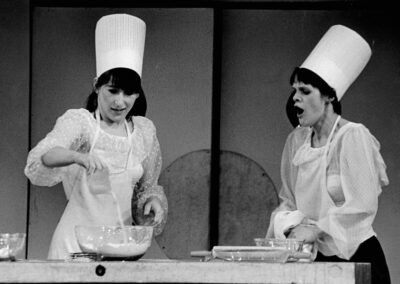 Two female chefs are baking, one barking instructions, the other pouring ingredients