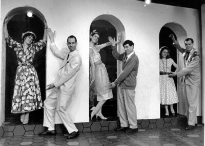 3 men and 3 women pose in front of a building. The women are positioned between 3 arches and each of the men are gesturing at each of the women