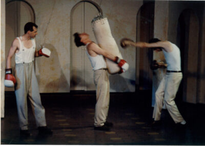 3 Men stand wearing boxing gloves. The man on the right punches a bag, while the man in the middle holds the bag and the man on the left looks on.