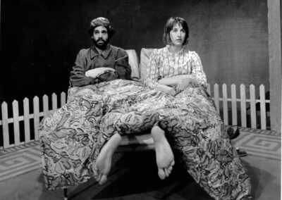 A man and woman are sitting upright in bed looking in opposite directions, tired. Another pair of feet pop out from the bottom of the blanket