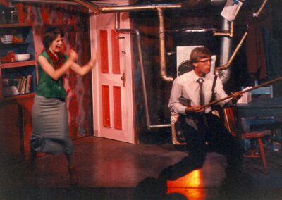 A man dressed in office clothes mimes playing a guitar on a walking stick, while a woman dressed for a evening looks on in glee