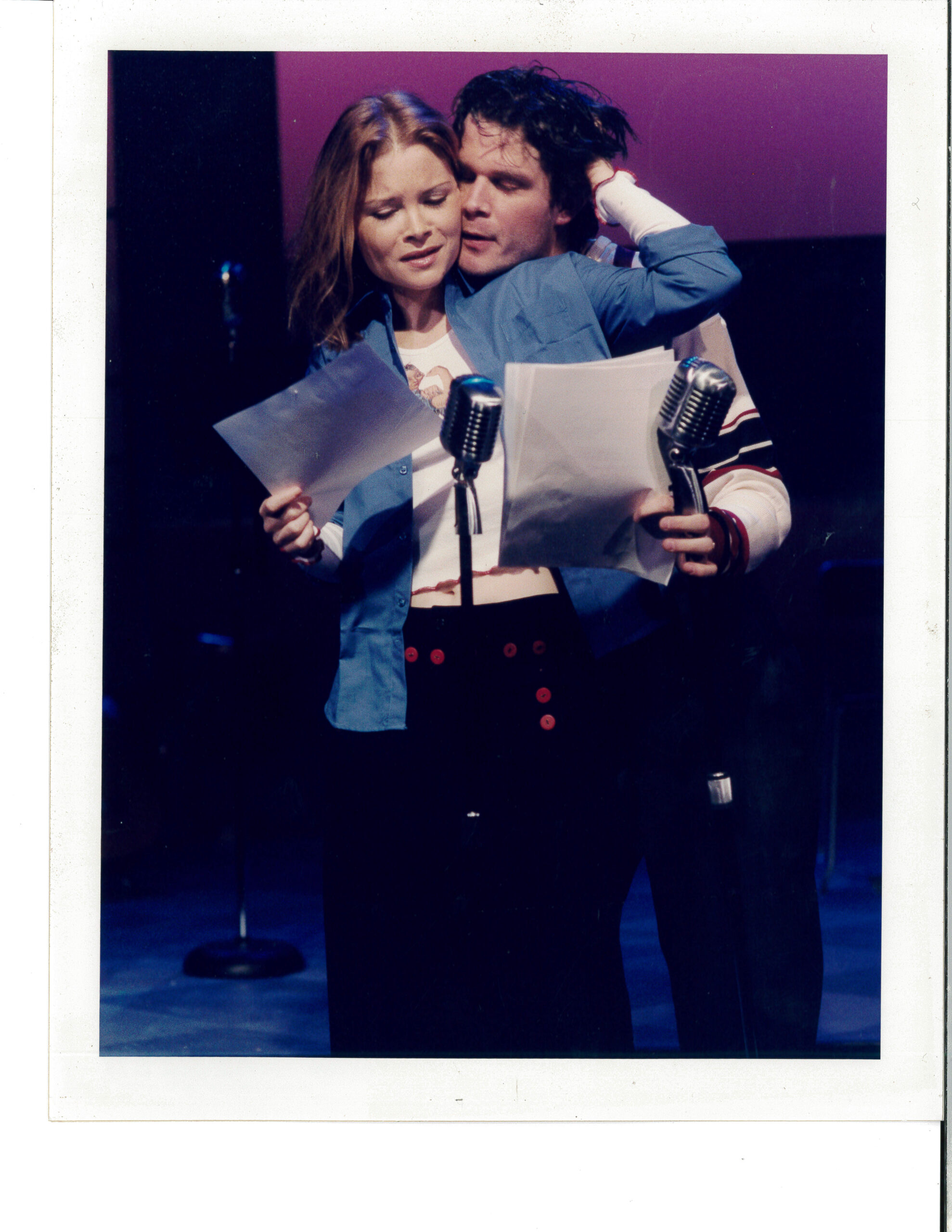 A man and woman hold each other lovingly while singing in front of radio mics, holding sheets of music