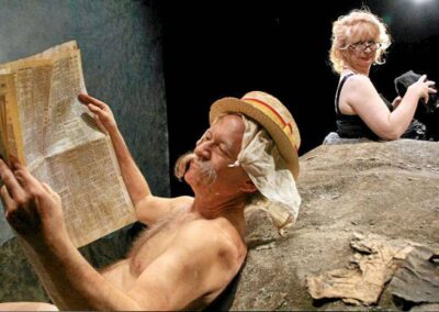 A blonde, middle-aged woman buried in sand wearing a black strappy dress looks over her right shoulder at a man whois reading a newspaper and wearing a straw hat