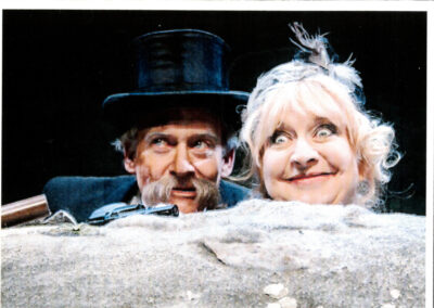 A middle aged couple, consisting of a blonde haired woman and a man in a top hat with a huge mustache peer out of the sand they're buried in. The woman looks manically happy, and the man looks off conspiratorially