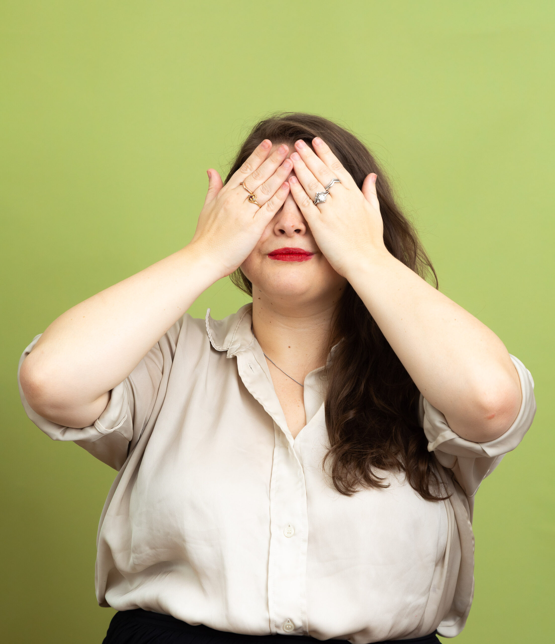Jess stands against a bright green background with both hands covering their eyes. They have long brown hair worn to one side and wear red lipstick. They are dressed in a light beige button-up shirt and wear rings on their fingers.