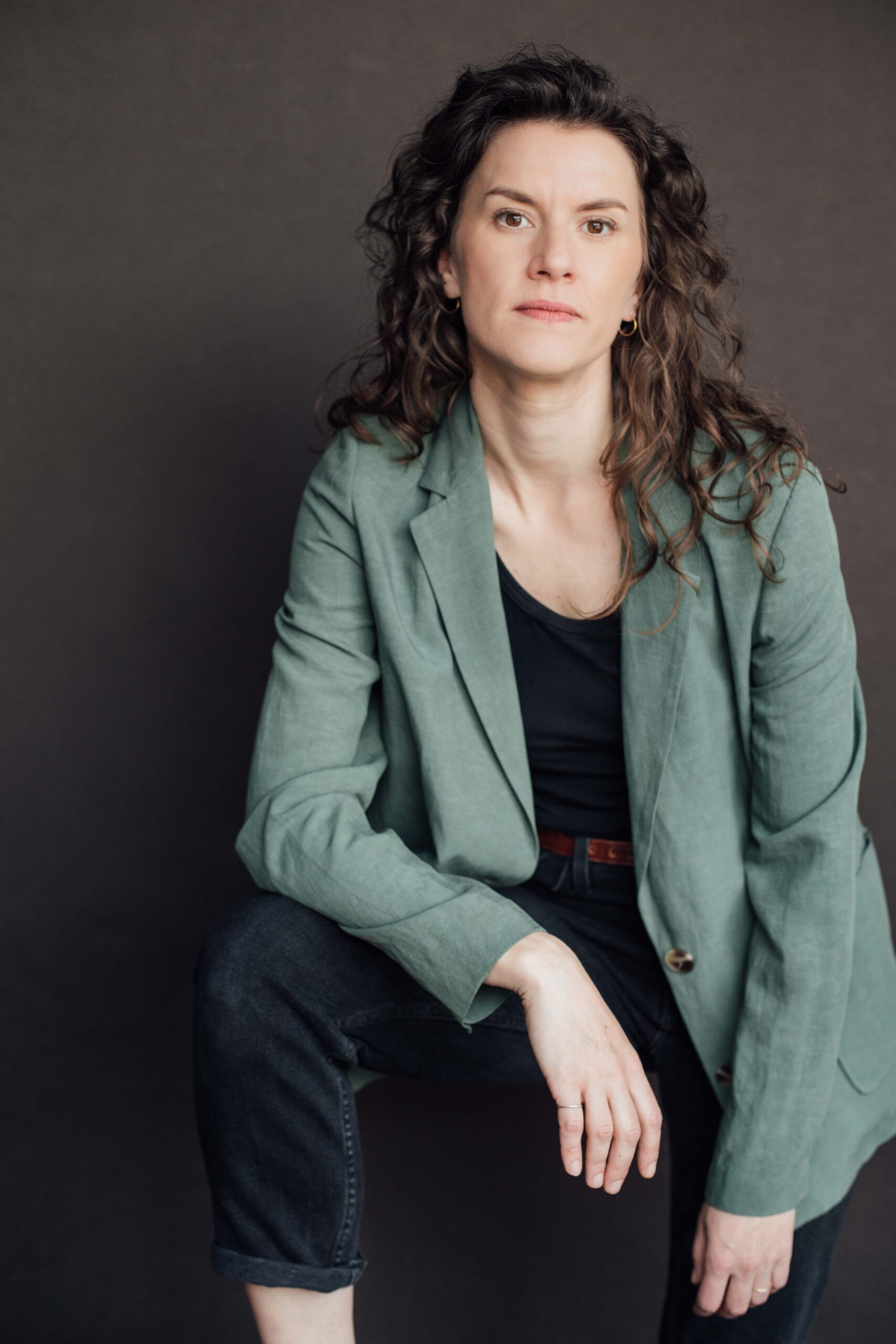 Headshot of Derek Kwan. He is an East Asian man in his 40s, with a shaved head and stubble moustache and beard. He wears a white shirt, a dark green corduroy vest, and a dark grey suit jacket adorned with a needle-felted pin of a fox head.
