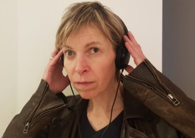 Alex Bulmer, a white woman with short dirty blond hair, wears a brown leather jacket and a black v neck top. They hold black over-ear headphones with both hands, and press them lightly against their ears. Their expression is focused and attentive - they look directly at the camera. The background is a plain, light-coloured wall.
