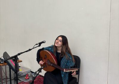 A young palestinian woman seated while holding an Oud. She is wearing a black dress and a turquoise patterned shawl. She is surrounded by several mics and a music stand. The floor is a patterned red rug.
