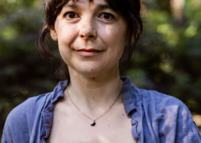 A young white woman smiles. She wears a grey strappy top and a blue shirt. She is standing in a leafy park.