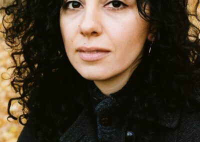 A palestinian woman with black hair in a black buttoned up jacked smiles quietly into the camera. She is surrounded by autumnal trees.