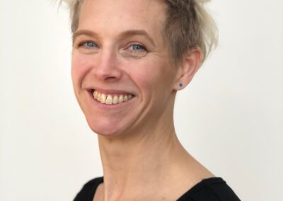 Sara stands smiling. She has short blonde hair and is wearing a black top. She is standing against a cream background.