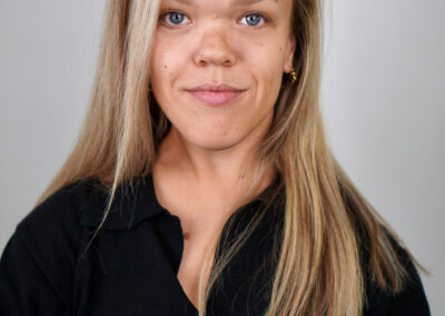 Alexia stands smiling with her mouth closed. She has long blond hair and is wearing a black top. The background is grey.