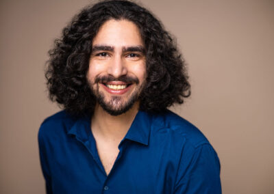 Yousef stands smiling in a deep blue shirt against a light brown background. He has shoulder length curly hair.